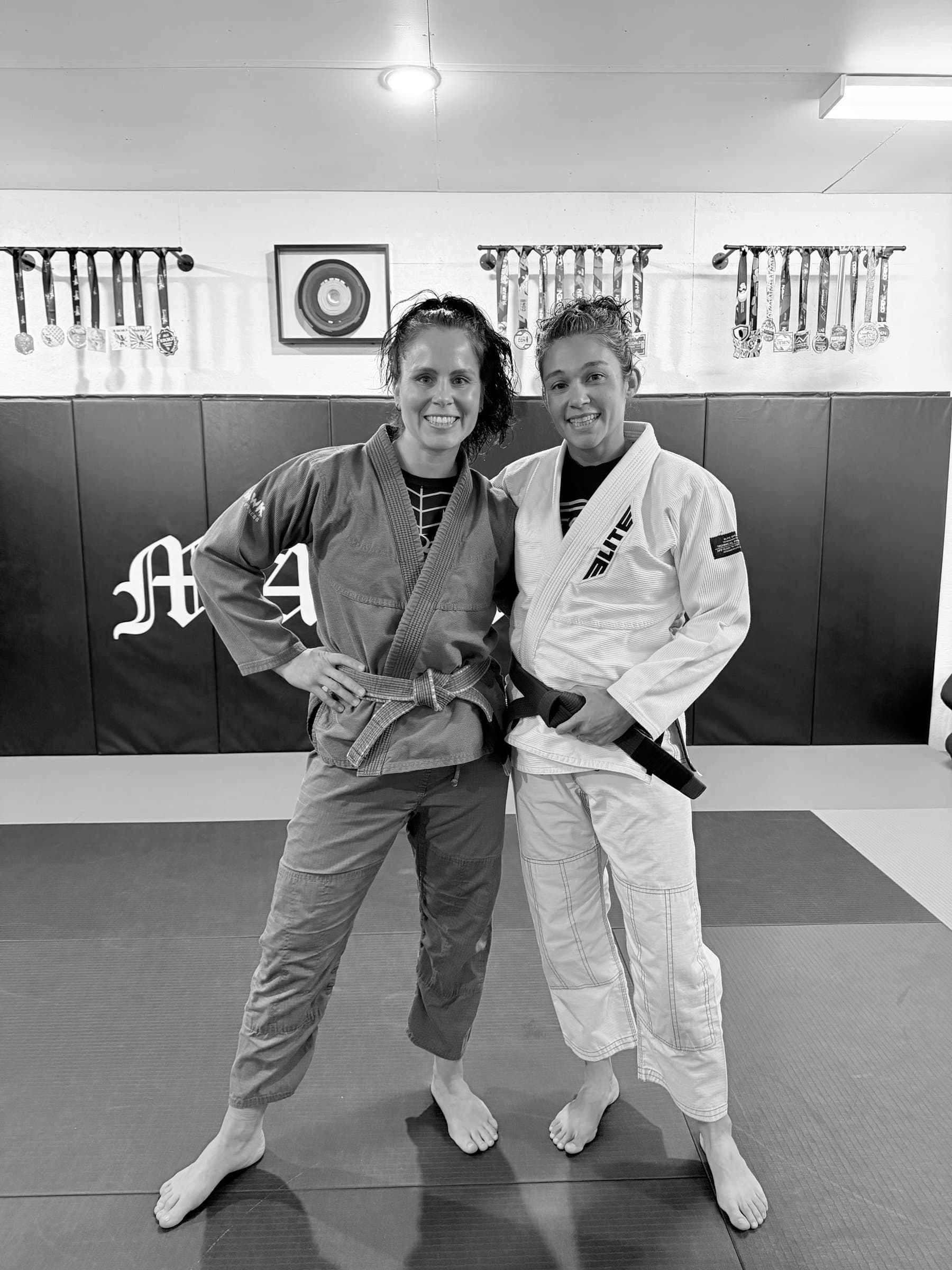 Two women in gis standing in a gym, smiling.